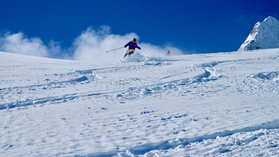 Ein Skifahrer springt über den Schnee in einem klaren blauen Himmel. Im Hintergrund sind schneebedeckte Berge zu sehen. | © Heli Rettensteiner