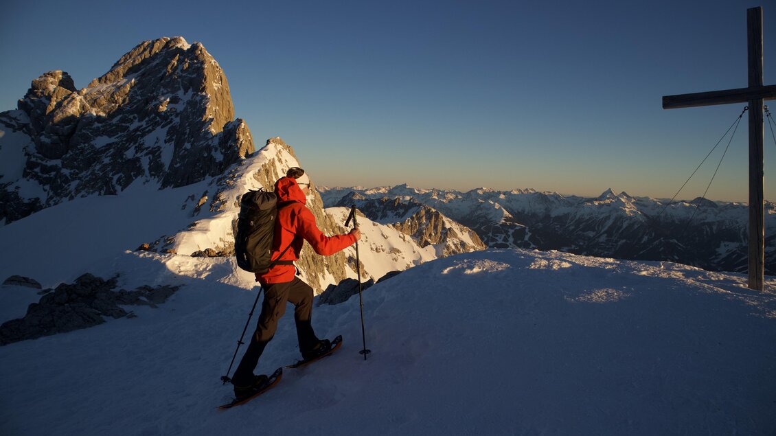 Ein Wanderer in roter Kleidung geht über verschneite Berge. Im Hintergrund sind majestätische Gipfel und ein Kreuz zu sehen. | © Heli Rettensteiner