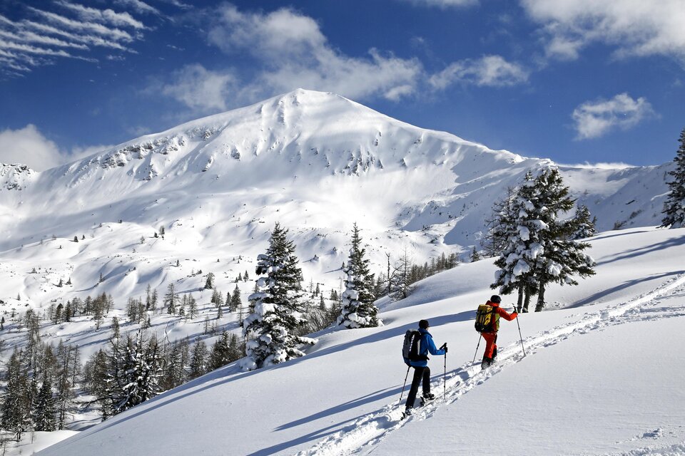Zwei Skifahrer erklimmen einen schneebedeckten Hang. Im Hintergrund erhebt sich ein majestätischer Berg unter einem blauen Himmel. | © Herbert Raffalt
