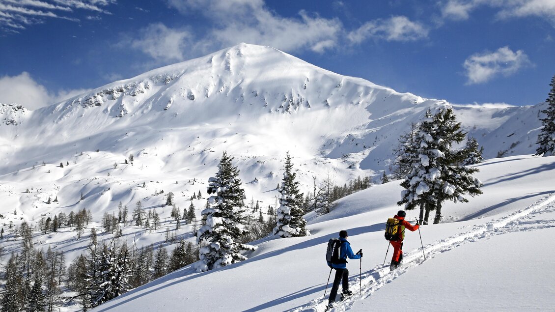 Zwei Skifahrer erklimmen einen schneebedeckten Hang. Im Hintergrund erhebt sich ein majestätischer Berg unter einem blauen Himmel. | © Herbert Raffalt