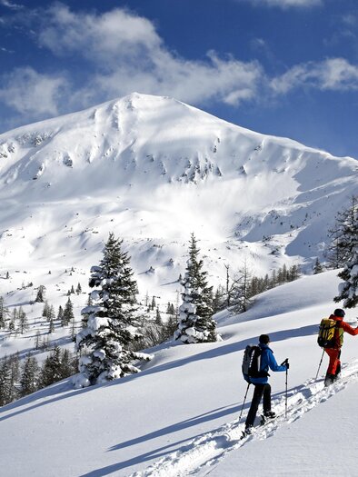 Two skiers ascend a snow-covered slope. In the background, a majestic mountain rises under a blue sky. | © Herbert Raffalt