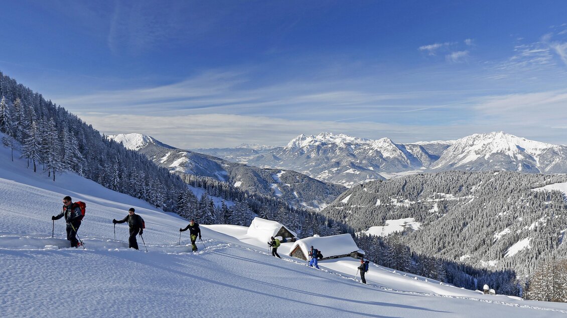 Eine Gruppe von Wanderern läuft durch die verschneite Landschaft in den Bergen. Im Hintergrund sind schneebedeckte Gipfel und ein klarer blauer Himmel sichtbar. | © Herbert Raffalt