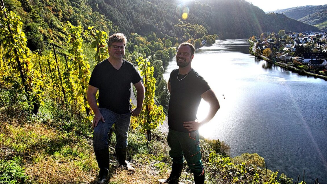 Zwei Männer stehen in einem Weinberg mit Blick auf einen Fluss. Im Hintergrund sind hügelige Landschaften und eine kleine Stadt zu sehen. | © Haberl & Fink