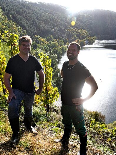 Two men are standing in a vineyard overlooking a river. In the background, hilly landscapes and a small town can be seen. | © Haberl & Fink