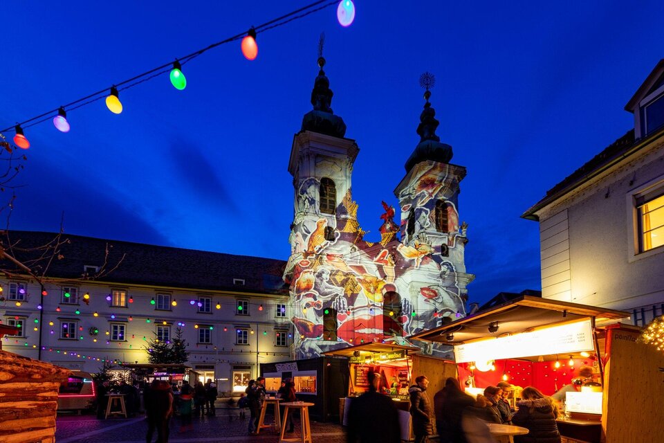 Ein schöner Weihnachtsmarkt bei Nacht mit bunten Lichtern. Im Hintergrund sind die beleuchteten Türme einer Kirche zu sehen. | © Graz Tourismus - Harry Schiffer