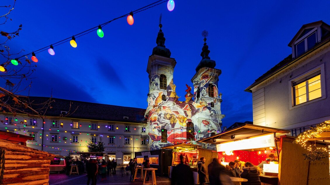 Ein schöner Weihnachtsmarkt bei Nacht mit bunten Lichtern. Im Hintergrund sind die beleuchteten Türme einer Kirche zu sehen. | © Graz Tourismus - Harry Schiffer