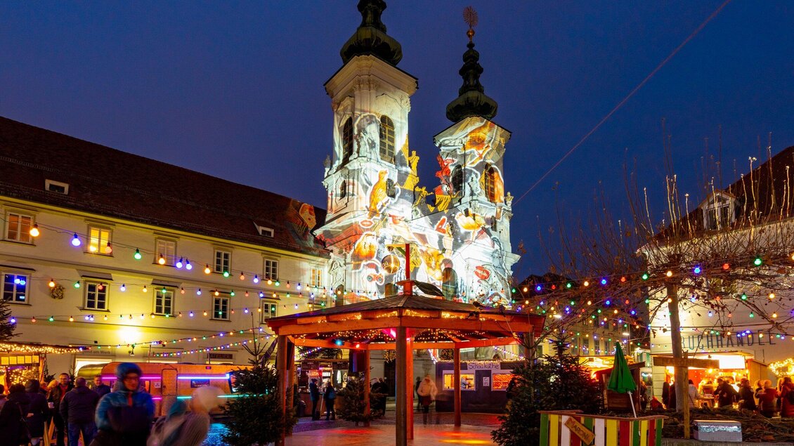 Ein festlich dekorierter Platz mit einem beleuchteten Kirchturm im Hintergrund. Viele Menschen genießen die Atmosphäre und die Weihnachtslichter. | © Graz Tourismus - Harry Schiffer