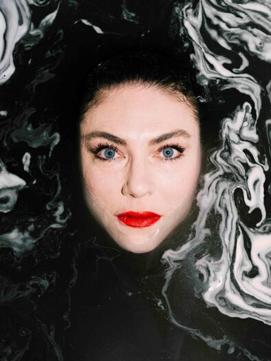 A woman with striking blue eyes and red lips stands in a dark-colored, frothy liquid. The contrast between her face and the background creates a fascinating atmosphere. | © Ingo Pertramer- Oper Graz