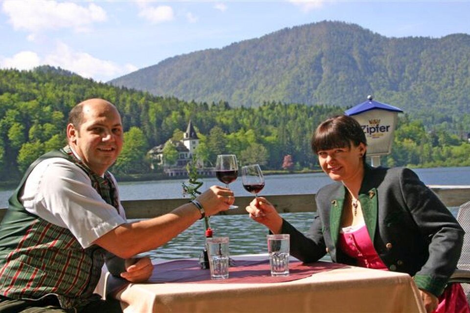 Ein Paar sitzt an einem Tisch am Wasser und prostet mit Rotwein. Im Hintergrund sind grüne Berge und ein schönes Anwesen zu sehen. | © Gasthaus Stöckl