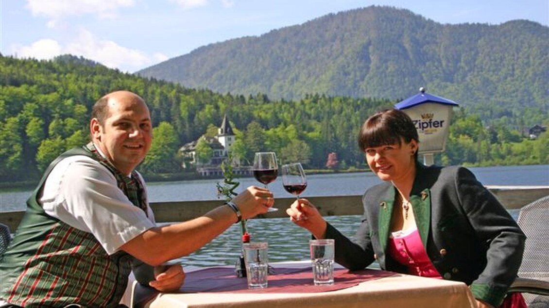 Ein Paar sitzt an einem Tisch am Wasser und prostet mit Rotwein. Im Hintergrund sind grüne Berge und ein schönes Anwesen zu sehen. | © Gasthaus Stöckl