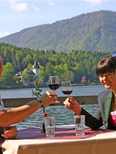 A couple sits at a table by the water, toasting with red wine. In the background, green mountains and a beautiful estate are visible. | © Gasthaus Stöckl