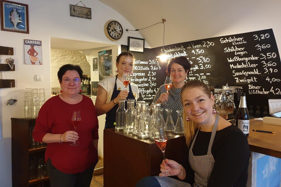Eine Gruppe von vier Frauen steht in einem gemütlichen Lokal und hält Gläser in der Hand. Im Hintergrund ist eine Tafel mit Getränkepreisen zu sehen. | © Weingut Resch