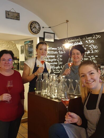 A group of four women is standing in a cozy establishment, holding glasses in their hands. In the background, a board with drink prices can be seen. | © Weingut Resch