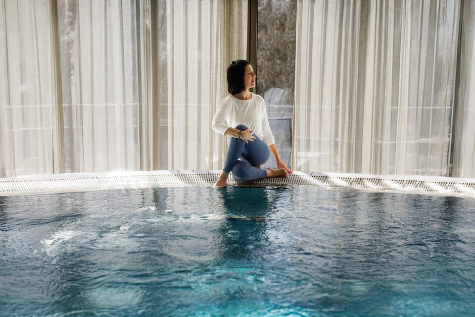 Eine Frau sitzt am Rand eines Innenpools und genießt die Atmosphäre. Weiche Vorhänge lassen sanftes Licht herein. | © ©Heilthermen Resort Bad Waltersdorf