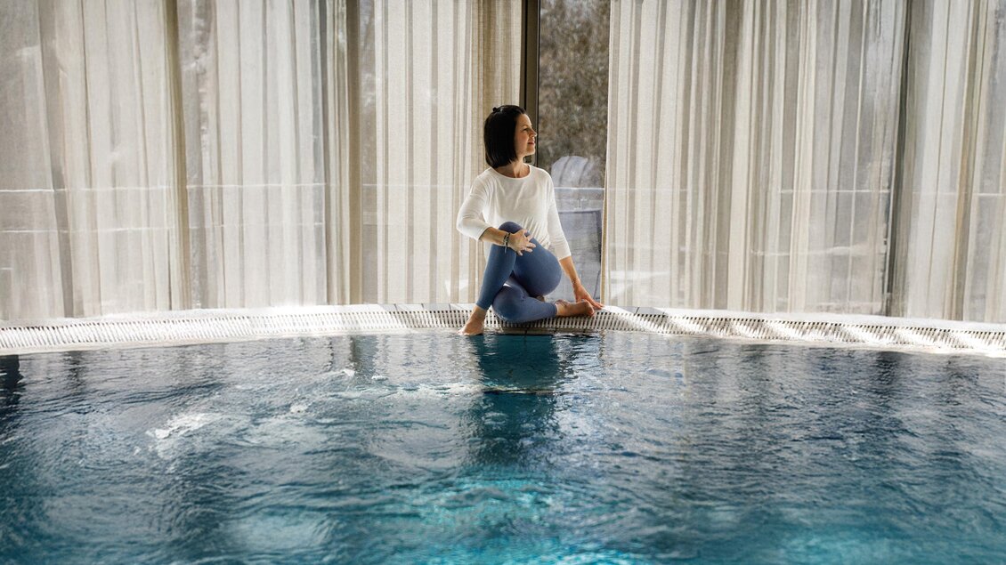 Eine Frau sitzt am Rand eines Innenpools und genießt die Atmosphäre. Weiche Vorhänge lassen sanftes Licht herein. | © ©Heilthermen Resort Bad Waltersdorf