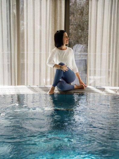 Eine Frau sitzt am Rand eines Innenpools und genießt die Atmosphäre. Weiche Vorhänge lassen sanftes Licht herein. | © ©Heilthermen Resort Bad Waltersdorf