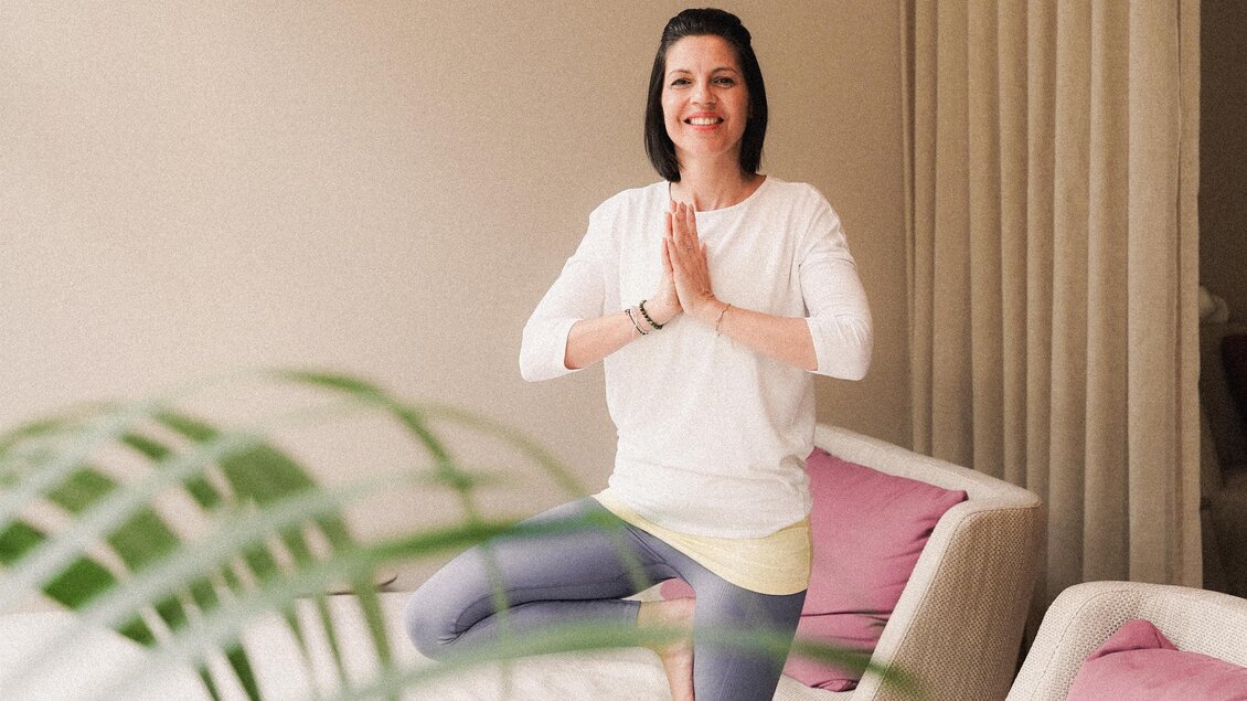 Eine Frau steht in einer Yoga-Pose und lächelt. Im Hintergrund sind helle Sofas mit Kissen und eine Pflanze sichtbar. | © Heilthermen Resort BW