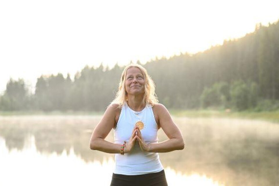 Eine Frau meditiert am Ufer eines ruhigen Sees. Sie hat die Hände vor der Brust gefaltet und lächelt in die Morgensonne.
