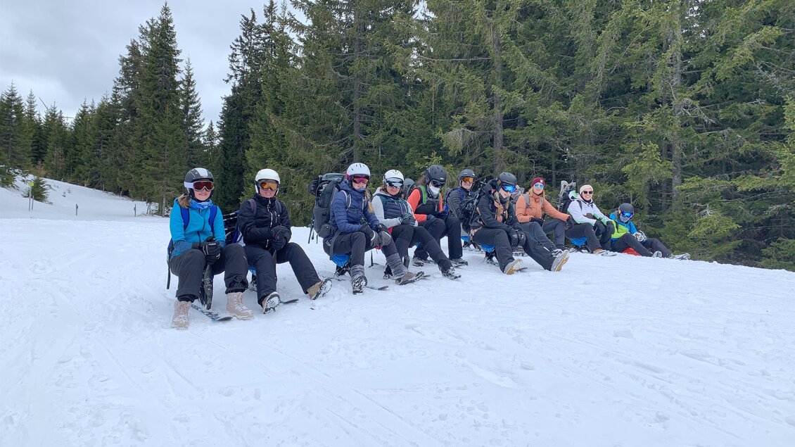 Eine Gruppe von Menschen sitzt auf einem schneebedeckten Berghang. Im Hintergrund sind Nadelbäume und eine winterliche Landschaft zu sehen. | © Steirisch Wandern