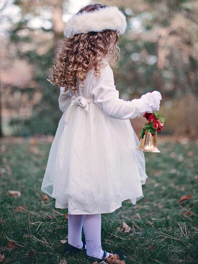 Ein kleines Mädchen steht mit dem Rücken zur Kamera. Es trägt ein weißes Kleid, eine helle Mütze und hält eine kleine Glocke sowie etwas Grünes in der Hand. | © Pixabay