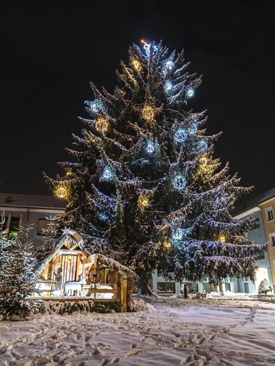 A large, festively lit Christmas tree stands in the snow. In the background, buildings decorated with lights are visible. | © Tourismus Murtal-Anita Fössl