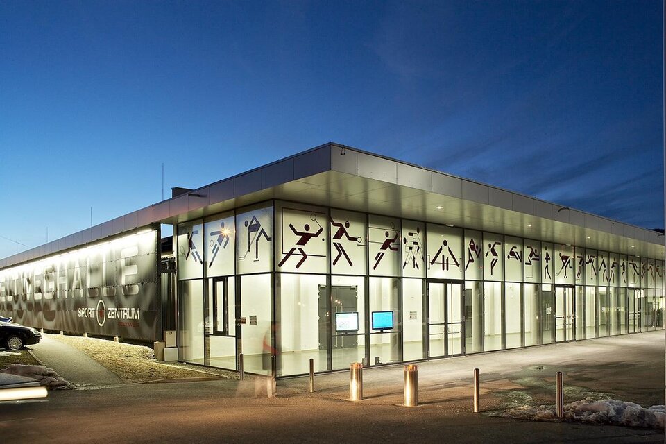 Ein modernes Sportzentrum mit großen Glasfenstern und sportlichen Motiven an der Fassade. Es ist Abend und das Gebäude wird von einem sanften Licht beleuchtet. | © Sportzentrum Zeltweg