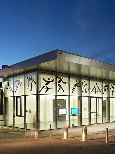 A modern sports center with large glass windows and athletic motifs on the facade. It is evening and the building is illuminated by a soft light. | © Sportzentrum Zeltweg