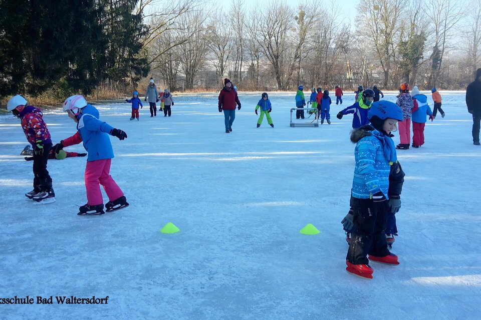 Eine Gruppe von Kindern läuft auf einem zugefrorenen See Schlittschuh. Im Hintergrund sind Erwachsene und weitere Kinder zu sehen, umgeben von schneebedeckten Bäumen. | © Volksschule Bad Waltersdorf