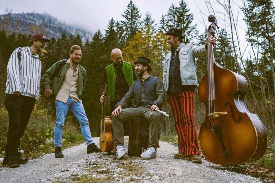 Eine Gruppe von fünf Musikern steht auf einem Weg unter Bäumen. Zwei Musiker haben Instrumente dabei, während die anderen entspannt miteinander interagieren. | © Philipp Egglmeier