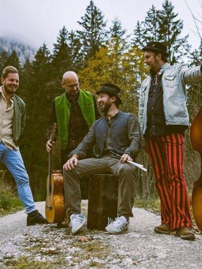 A group of five musicians stands on a path under trees. Two musicians have instruments with them, while the others are relaxing and interacting with each other. | © Philipp Egglmeier
