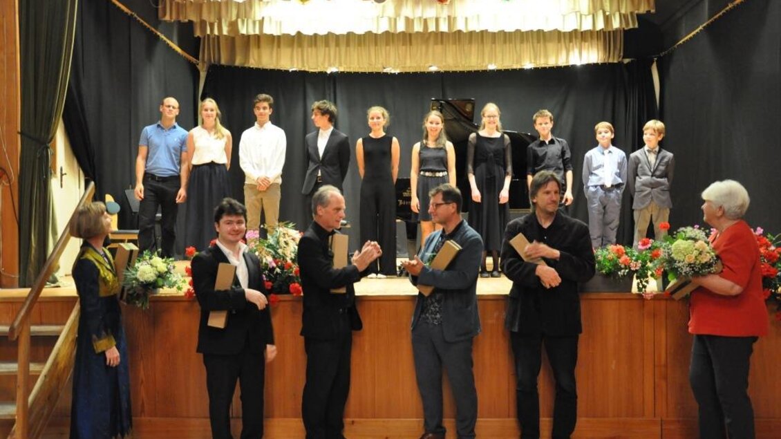 Eine Gruppe von jungen Menschen steht auf der Bühne, während einige Erwachsene im Vordergrund interagieren. Blumenarrangements und festliche Kleidung sind sichtbar, was auf eine besondere Veranstaltung hinweist. | © Kulturreferat der Marktgemeinde Irdning-Donnersbachtal