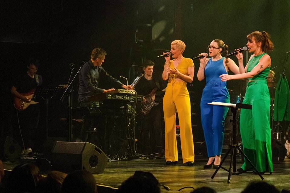 Ein Live-Konzert mit drei Sängerinnen in bunten Outfits, die auf der Bühne singen. Im Hintergrund spielt eine Band Musik. | © Helena Melbinger