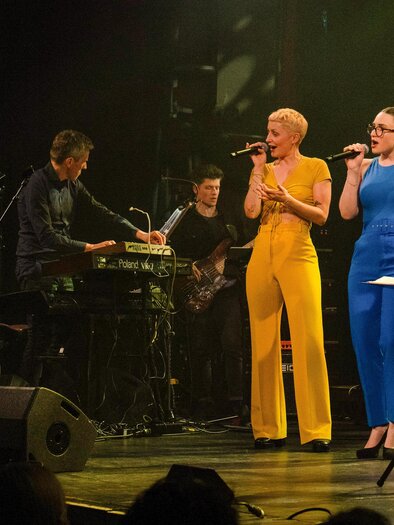 Ein Live-Konzert mit drei Sängerinnen in bunten Outfits, die auf der Bühne singen. Im Hintergrund spielt eine Band Musik. | © Helena Melbinger
