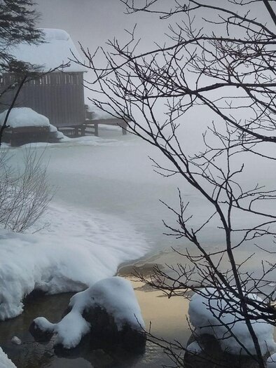 Eine winterliche Landschaft mit einem gefrorenen See und Schnee. Im Hintergrund steht eine Holzstr Struktur, umgeben von nebliger Atmosphäre. | © Christina Arzberger