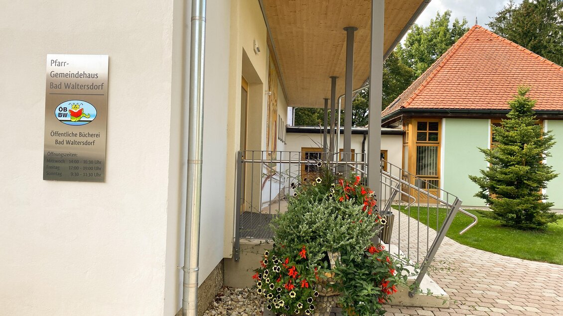 Ein Gebäude mit einer modernen Fassade und einem gepflegten Eingangsbereich. Bunte Pflanzen schmücken den Eingangsbereich und eine Terrasse ist sichtbar. | © Thermen- und Vulkanland