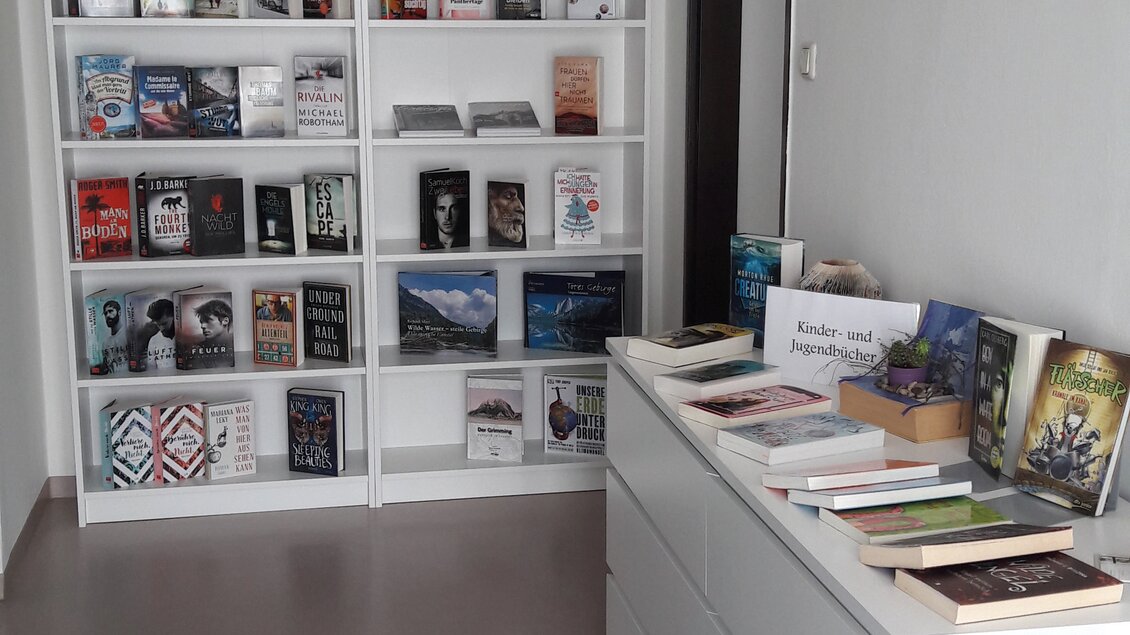 Ein modernes Bücherregal mit verschiedenen Büchern steht an der Wand. Daneben befindet sich eine schlichte Kommode mit weiteren Büchern darauf. | © Bibliothek der MG Stainach-Pürgg