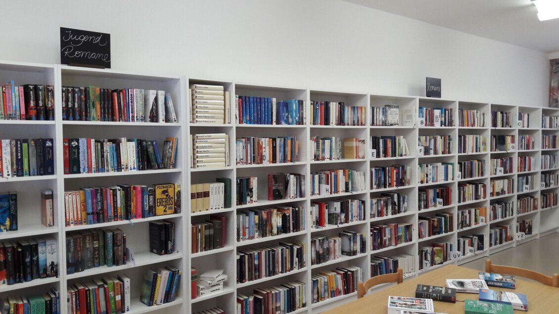 Ein Raum mit mehreren Bücherregalen, die voll mit verschiedenen Büchern sind. Auf einem Regal steht ein Schild mit der Aufschrift "Vorgeschlagene Romane". | © Bibliothek der MG Stainach-Pürgg