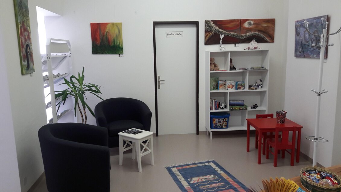 Ein gemütlicher Wartebereich mit zwei schwarzen Sessel und einem kleinen Tisch. An den Wänden hängen farbige Bilder, und ein Regal bietet Bücher und Spielsachen. | © Bibliothek der MG Stainach-Pürgg