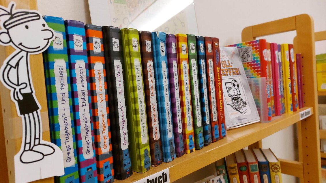 Eine Bücherregal mit bunten Kinderbüchern steht im Vordergrund. Auf der linken Seite ist eine Figur aus einem Comics zu sehen. | © Bibliothek der MG Stainach-Pürgg