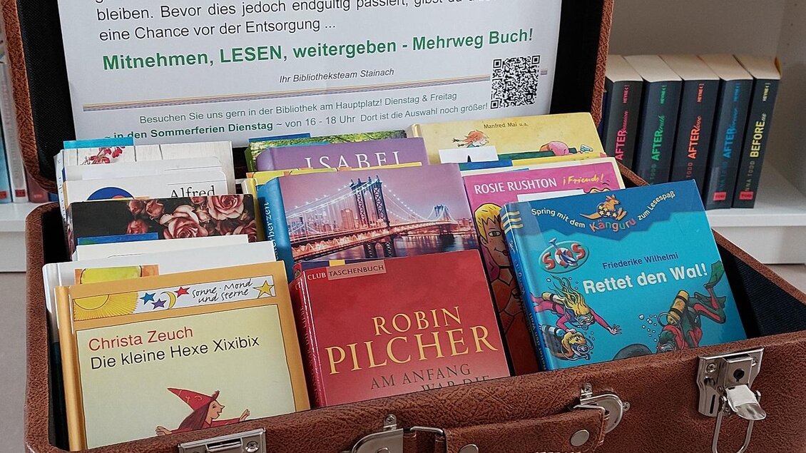 Eine offene Kiste mit verschiedenen Büchern steht auf einem Tisch. Im Hintergrund ist ein Schild mit Informationen über die Bibliothek und eine Aufforderung zum Lesen zu sehen. | © ÖB der MG Stainach-Pürgg