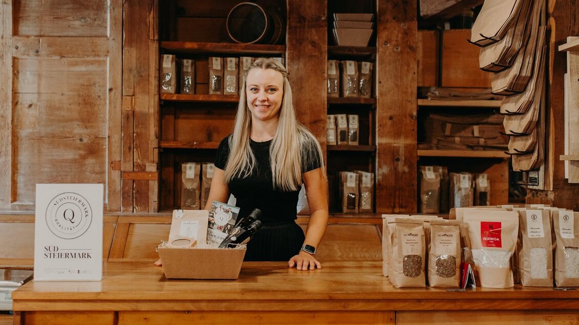 Eine freundliche Frau steht hinter einem Holztresen in einem Geschäft. Im Hintergrund sind verschiedene Verpackungen und Produkte sichtbar. | © Regionalmanagement Südweststeiermark GmbH/webquartier