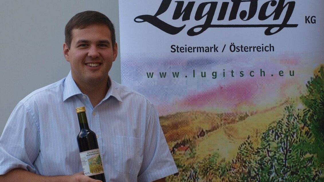 Ein lächelnder Mann hält eine Flasche Öl in der Hand. Im Hintergrund ist ein Banner der Ölmühle Lugitsch zu sehen. | © Ölmühle Lugitsch