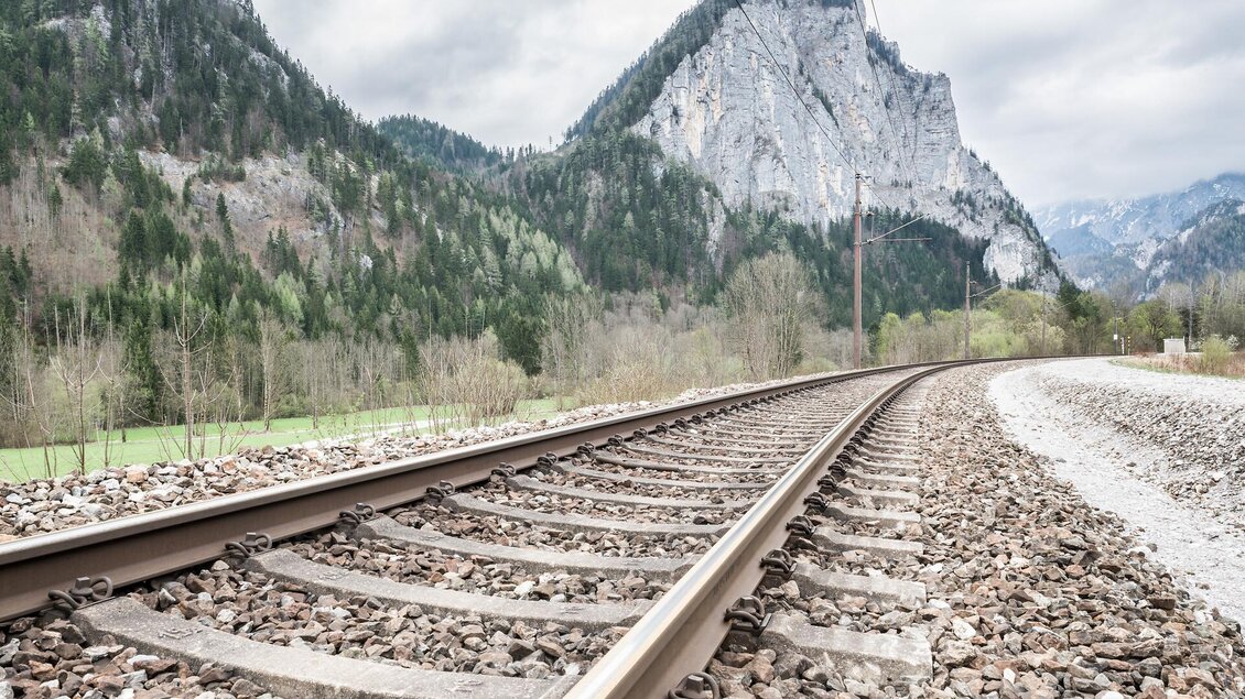 Ein Schienenweg verläuft durch eine malerische Landschaft mit Bergen im Hintergrund. Die Wolken am Himmel sorgen für eine stimmungsvolle Atmosphäre.