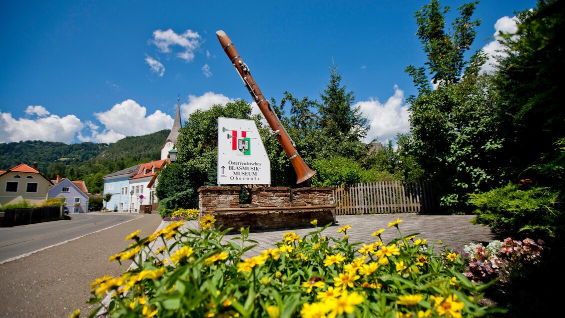 Ein großer, rustikaler Außenschild mit einer Trompete steht in einer malerischen Landschaft. Umgeben von bunten Blumen und grünen Bäumen unter einem blauen Himmel. | © Holzwelt Murau