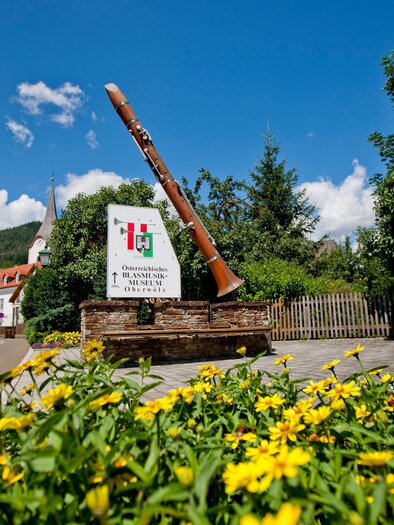Ein großer, rustikaler Außenschild mit einer Trompete steht in einer malerischen Landschaft. Umgeben von bunten Blumen und grünen Bäumen unter einem blauen Himmel. | © Holzwelt Murau