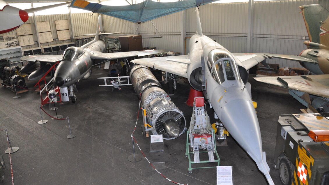 Zwei Militärflugzeuge in einem Hangar, eines mit einem konventionellen Design und das andere mit einem futuristischen Aussehen. Neben den Flugzeugen sind verschiedene technische Geräte und ein Triebwerk zu sehen. | © Österreichisches Luftfahrtmuseum