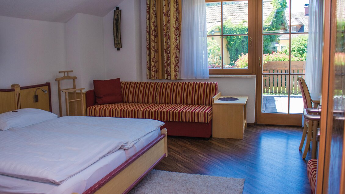 Ein helles Zimmer mit einem Doppelbett und einer gemütlichen Couch. Große Fenster bieten einen Blick auf den Garten. | © Gerhard Schernthaner