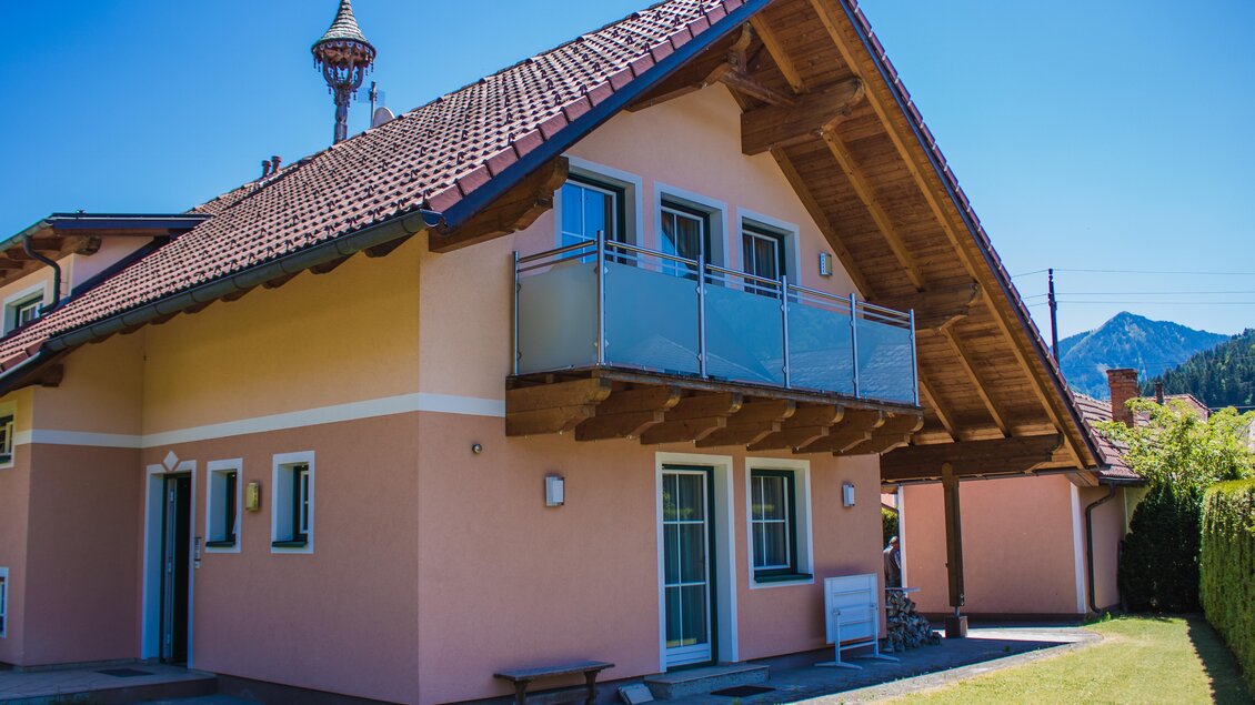 Ein modernes, pinkfarbenes Haus mit einem großen Dach und einem Balkon. Im Hintergrund sind Berge und ein klarer blauer Himmel zu sehen. | © Gerhard Schernthaner