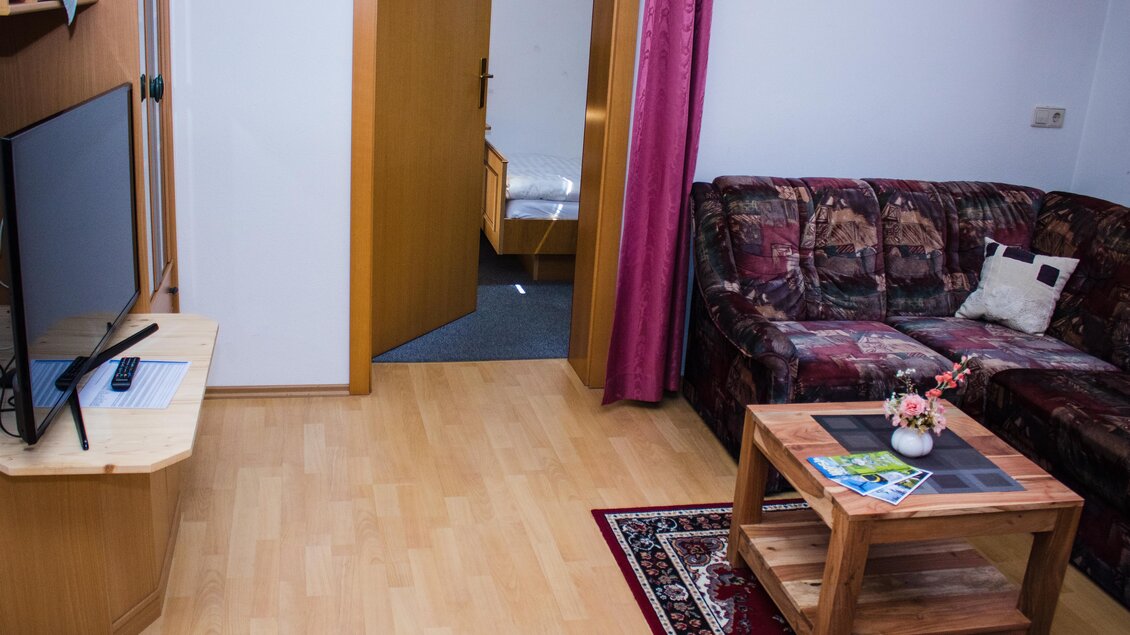 Ein gemütlicher Wohnbereich mit einem Sofa, einem Fernseher und einem Holzkaffeetisch. Im Hintergrund ist ein offenes Schlafzimmer zu sehen. | © Gerhard Schernthaner