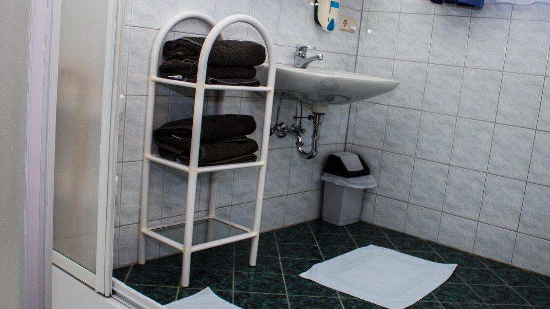 Ein modernes Badezimmer mit einem Waschbecken, einer Dusche und einem Handtuchregal. Auf dem Boden liegen zwei Handtücher und ein Mülleimer ist sichtbar. | © Gerhard Schernthaner